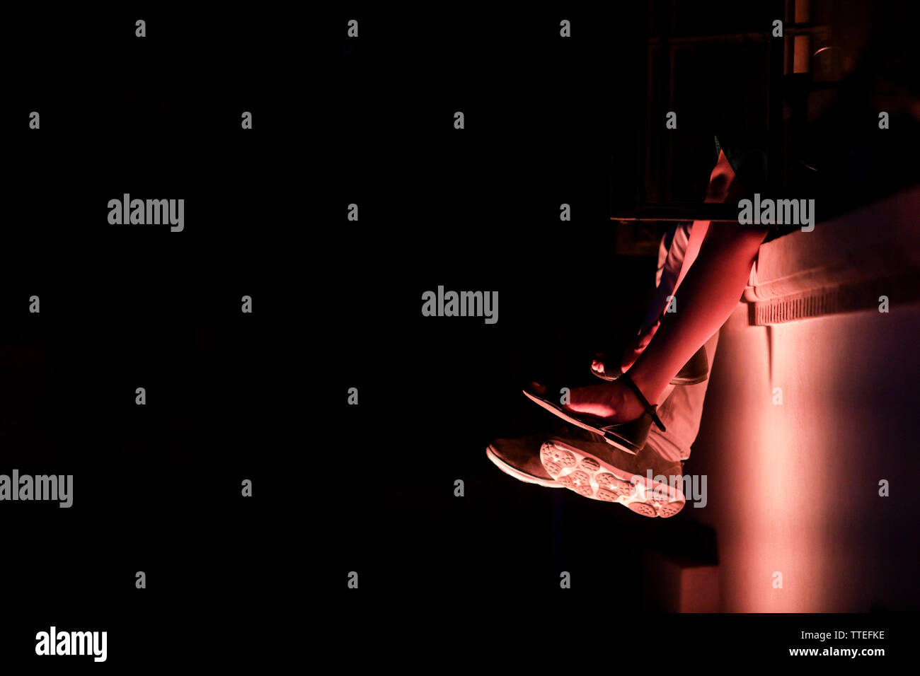 Dettagli con i piedi di una giovane coppia (i suoi sandali e il suo sneakers), seduto su di una mensola di pietra, di notte e con una forte luce contrastanti. Foto Stock