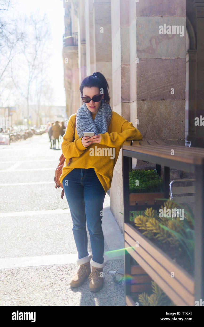 Moda turista femminile di indossare un abbigliamento caldo utilizzando smart phone mentre in piedi in città Foto Stock