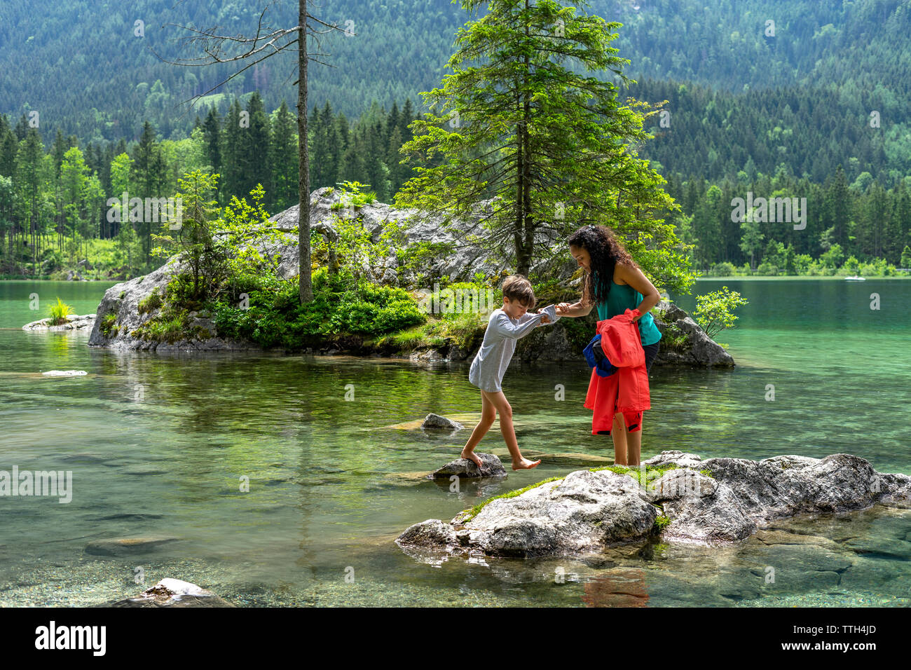 Madre aiutando il figlio a saltare da una roccia a un altro in un lago Foto Stock