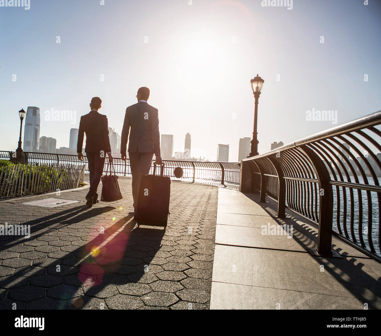 Vista posteriore di imprenditori a piedi con i bagagli mentre passeggiate sul lungomare durante la giornata di sole Foto Stock