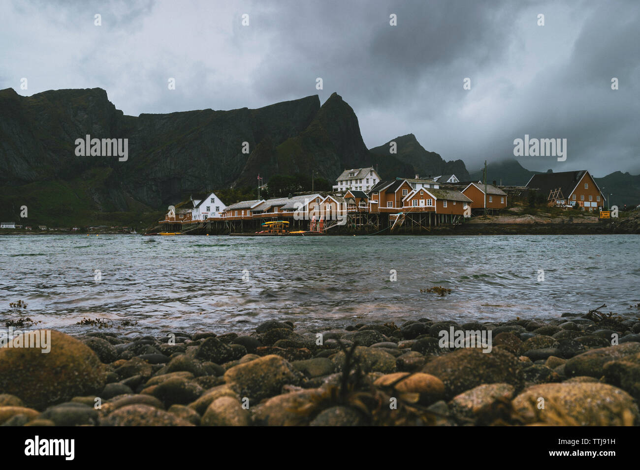 Case Lofoten catturato dalla riva di pietre con le montagne sullo sfondo dell'immagine. Giorno nuvoloso, mare calmo, orizzontale, sensazione di coldn Foto Stock