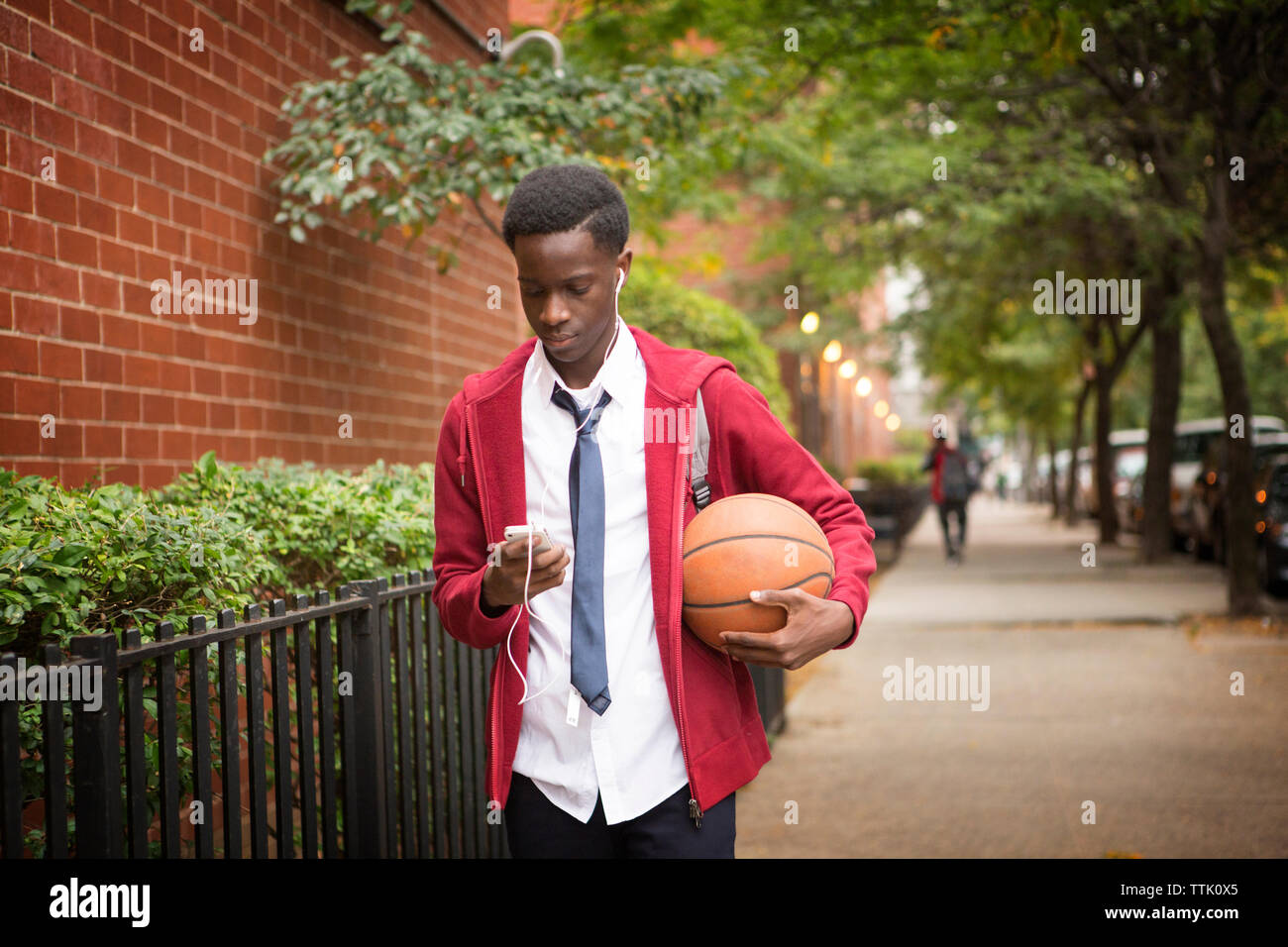 studente che usa il telefono e tiene il basket mentre cammina sul marciapiede Foto Stock