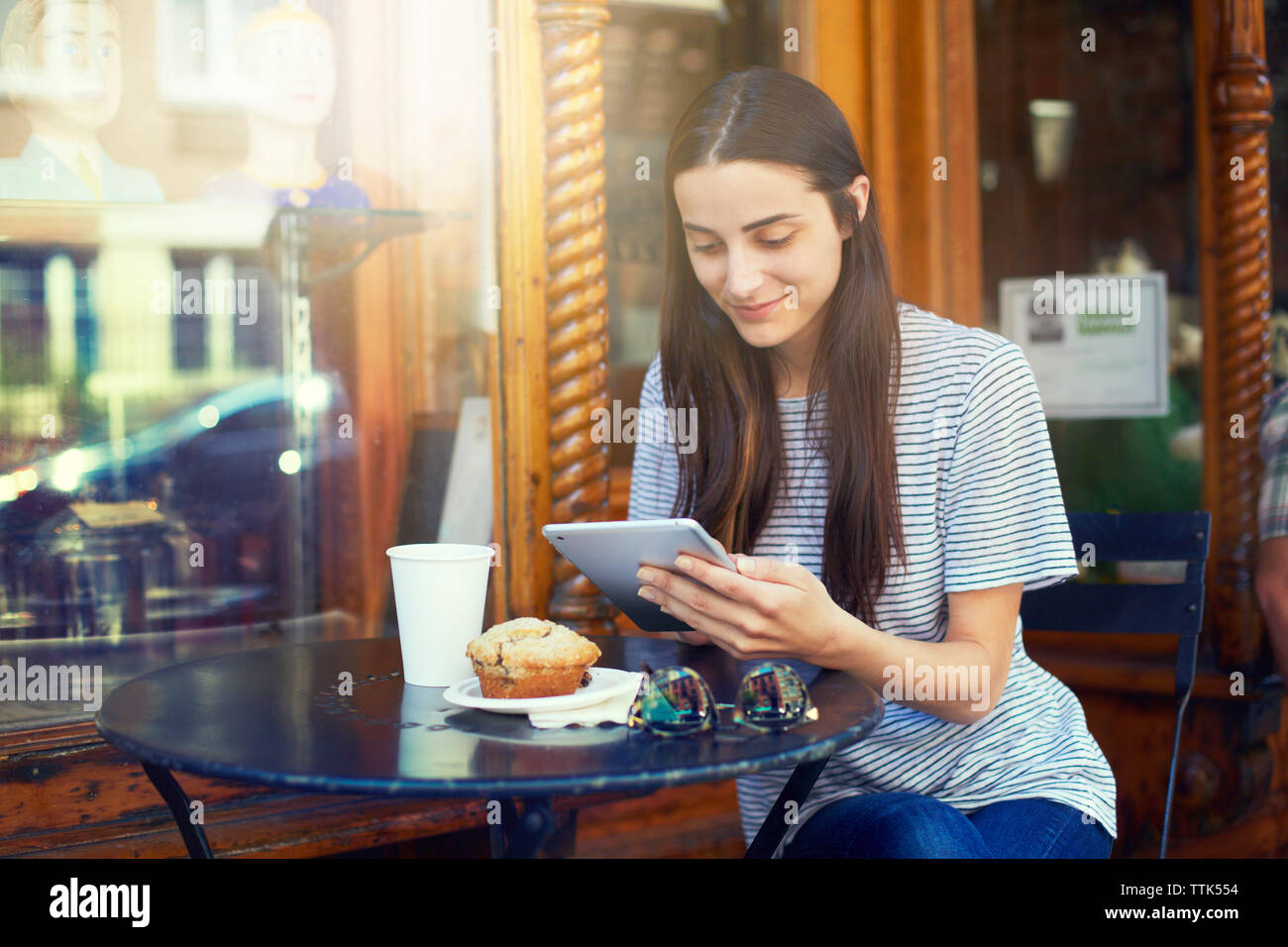 Donna sorridente con tavoletta digitale mentre seduta al tavolo al cafè sul marciapiede Foto Stock