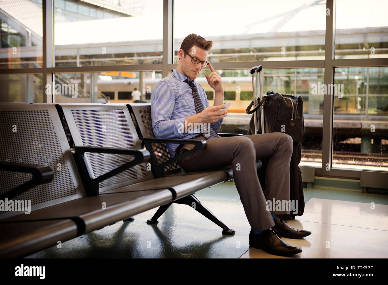 uomo d'affari che usa lo smartphone mentre siede seduto nella sala d'attesa della stazione ferroviaria Foto Stock