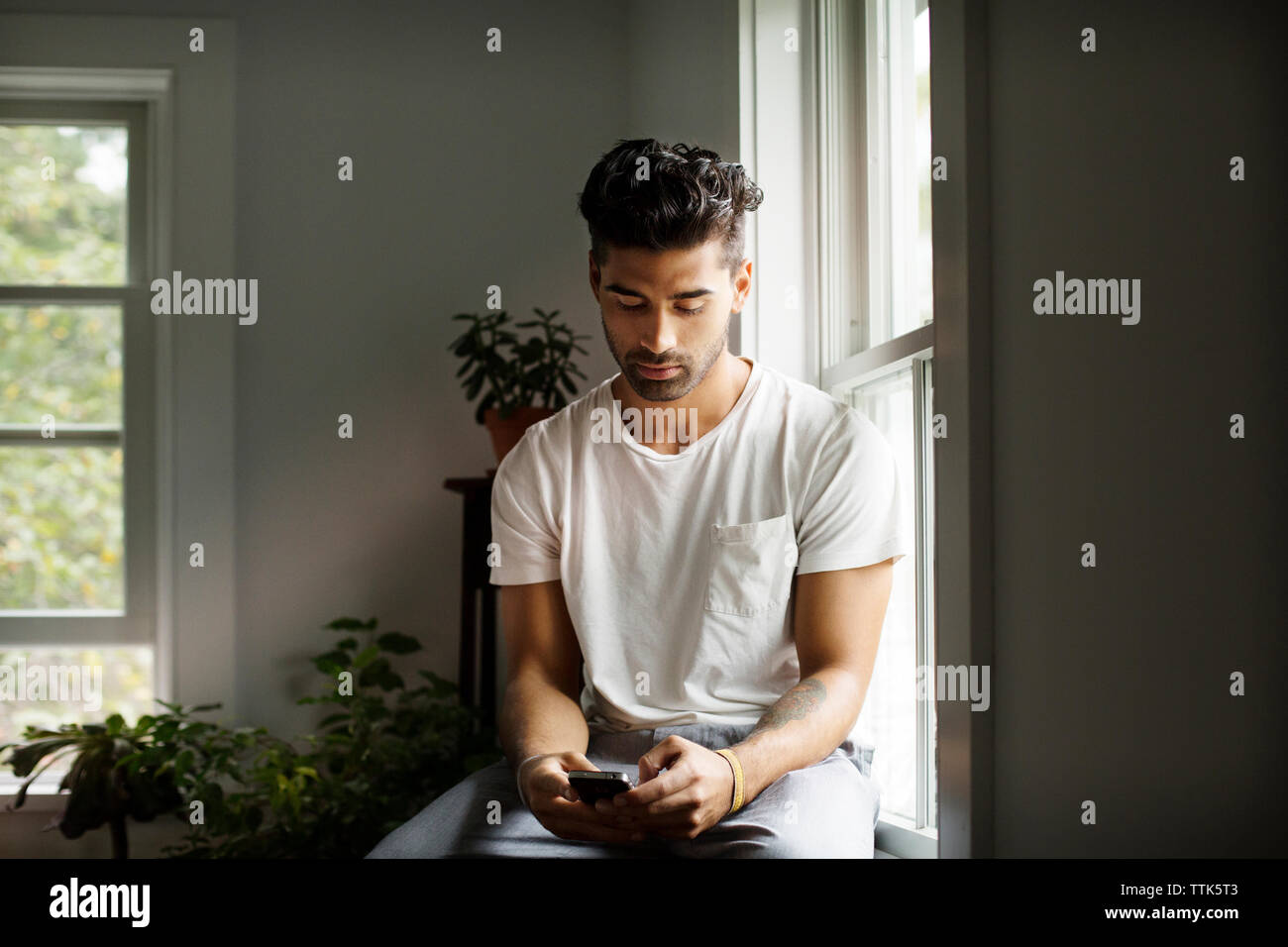 Giovane uomo utilizzando smart phone seduti dalla finestra a casa Foto Stock