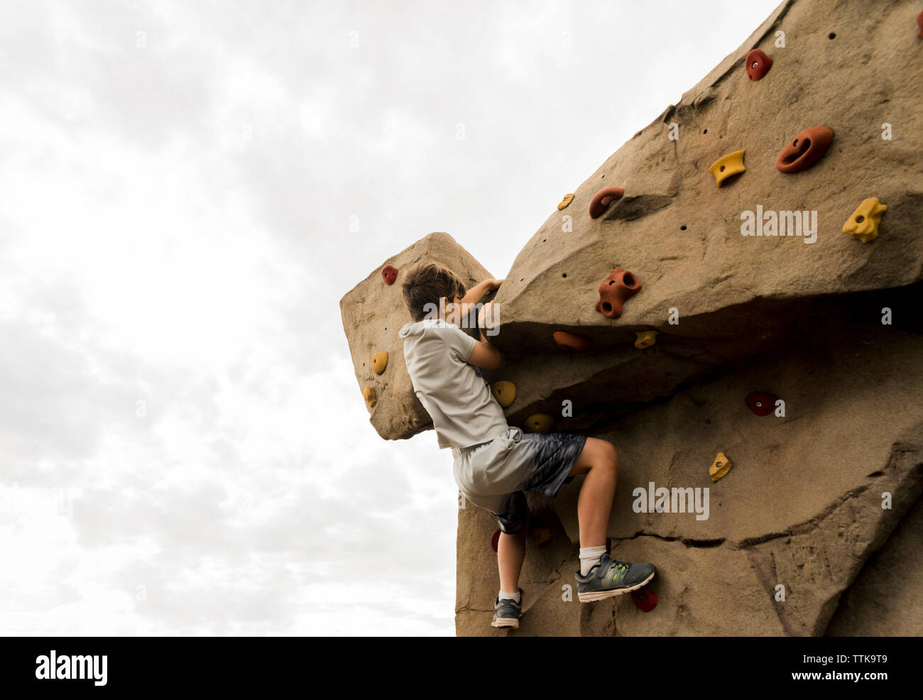 Basso angolo vista del ragazzo arrampicata boulder contro il cielo nuvoloso al parco giochi Foto Stock