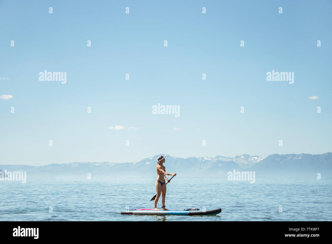 Vista laterale della donna in bikini paddleboarding sul lago contro il cielo blu durante la giornata di sole Foto Stock