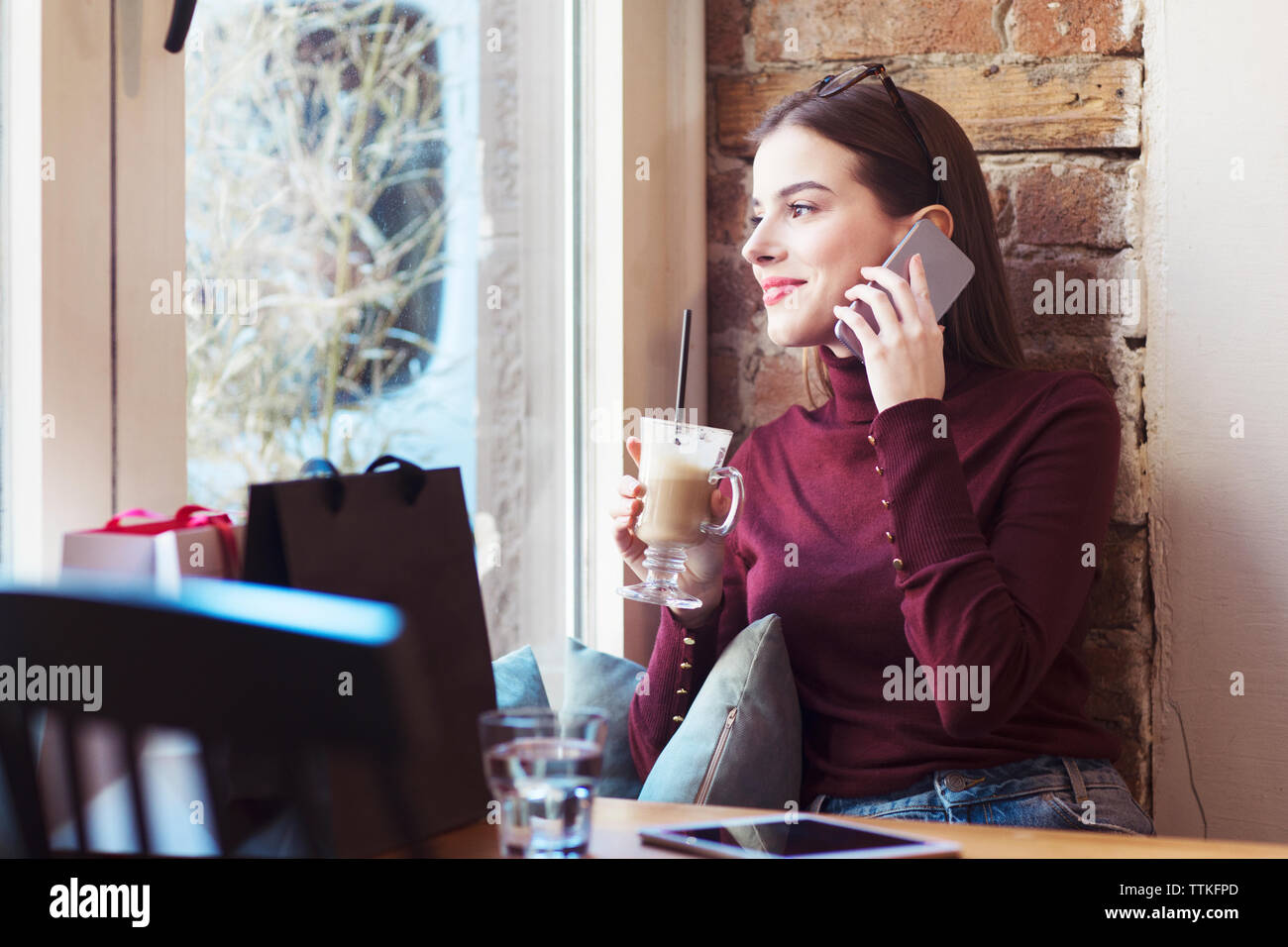 Donna con drink guardando attraverso la finestra mentre si parla al telefono cellulare in cafe Foto Stock