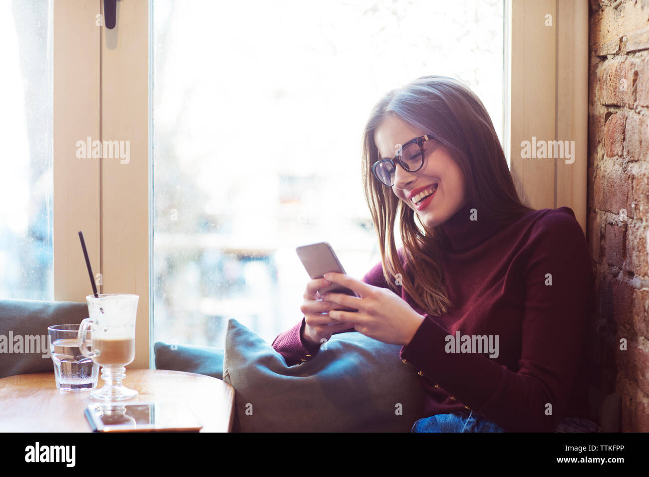 Donna felice usando il cellulare mentre seduta dalla finestra nel cafe Foto Stock