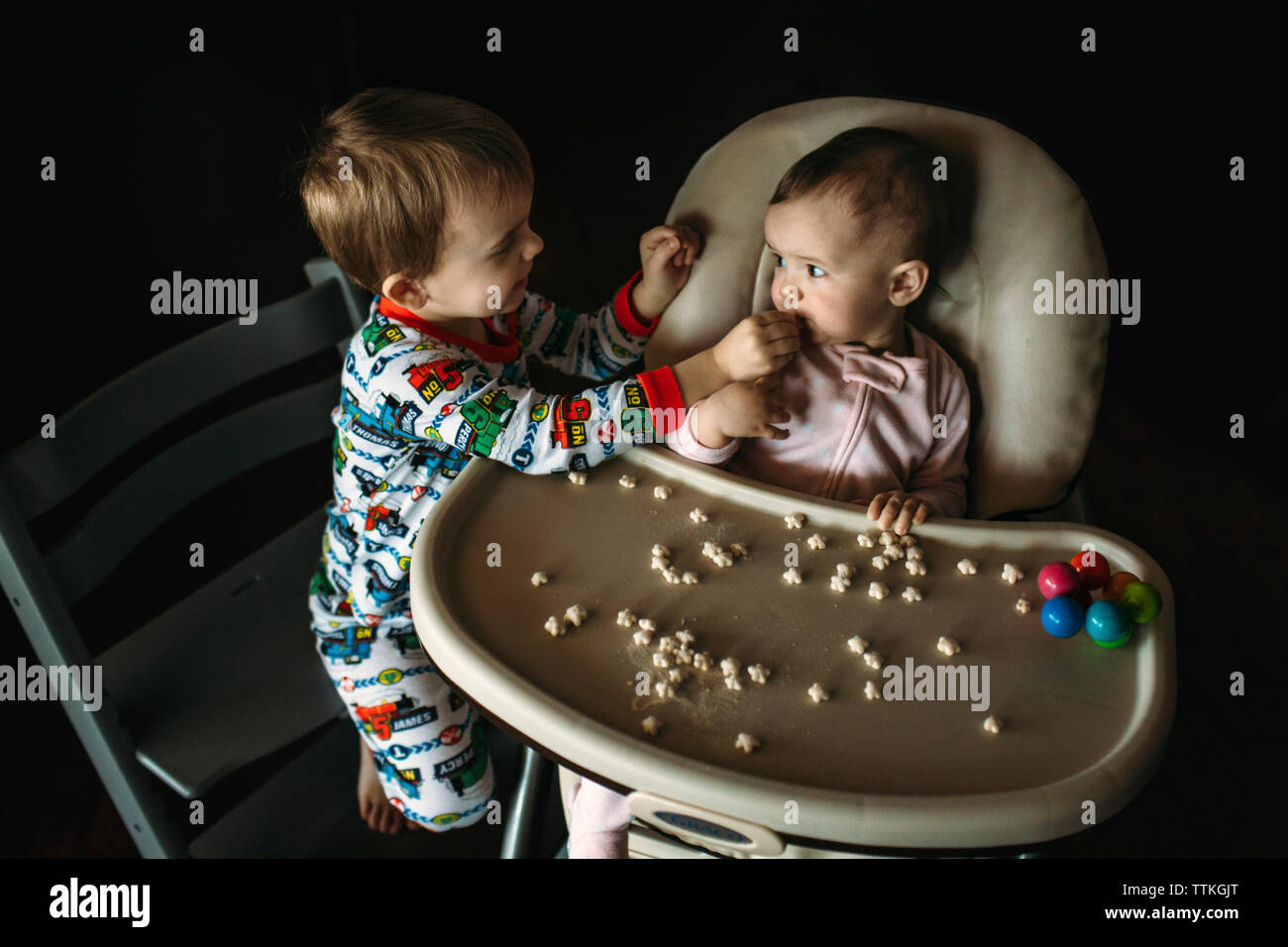 Fratello alimentare alimenti per neonati a suor seduto sulla sedia alta in camera oscura a casa Foto Stock