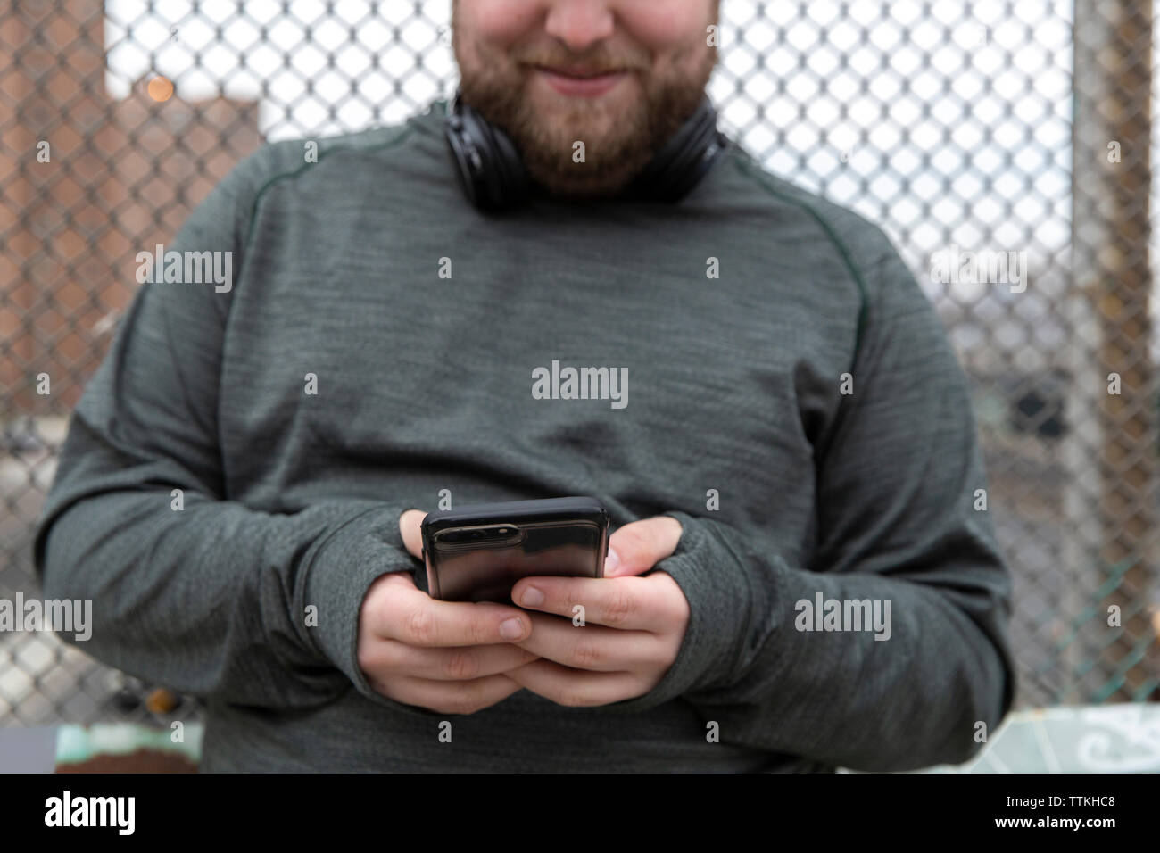 Sezione mediana del sovrappeso uomo utilizzando smart phone mentre in piedi contro la recinzione in città Foto Stock