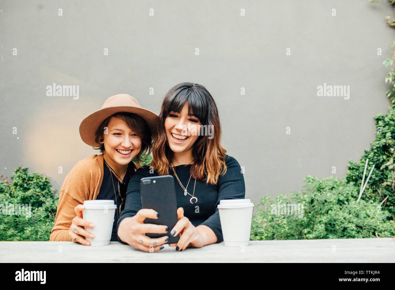 Donna sorridente amici prendendo selfie seduti a tavola contro la parete Foto Stock