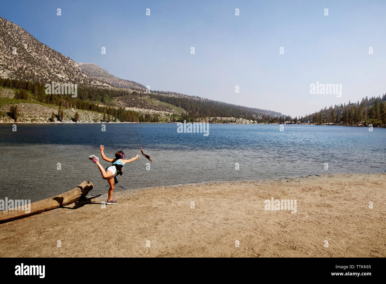 Ragazza in piedi su una gamba sola a riverbank contro le montagne e il cielo chiaro Foto Stock