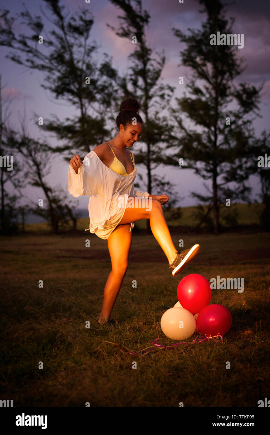 Vista laterale di felice adolescente scoppiare palloncini in campo al crepuscolo Foto Stock