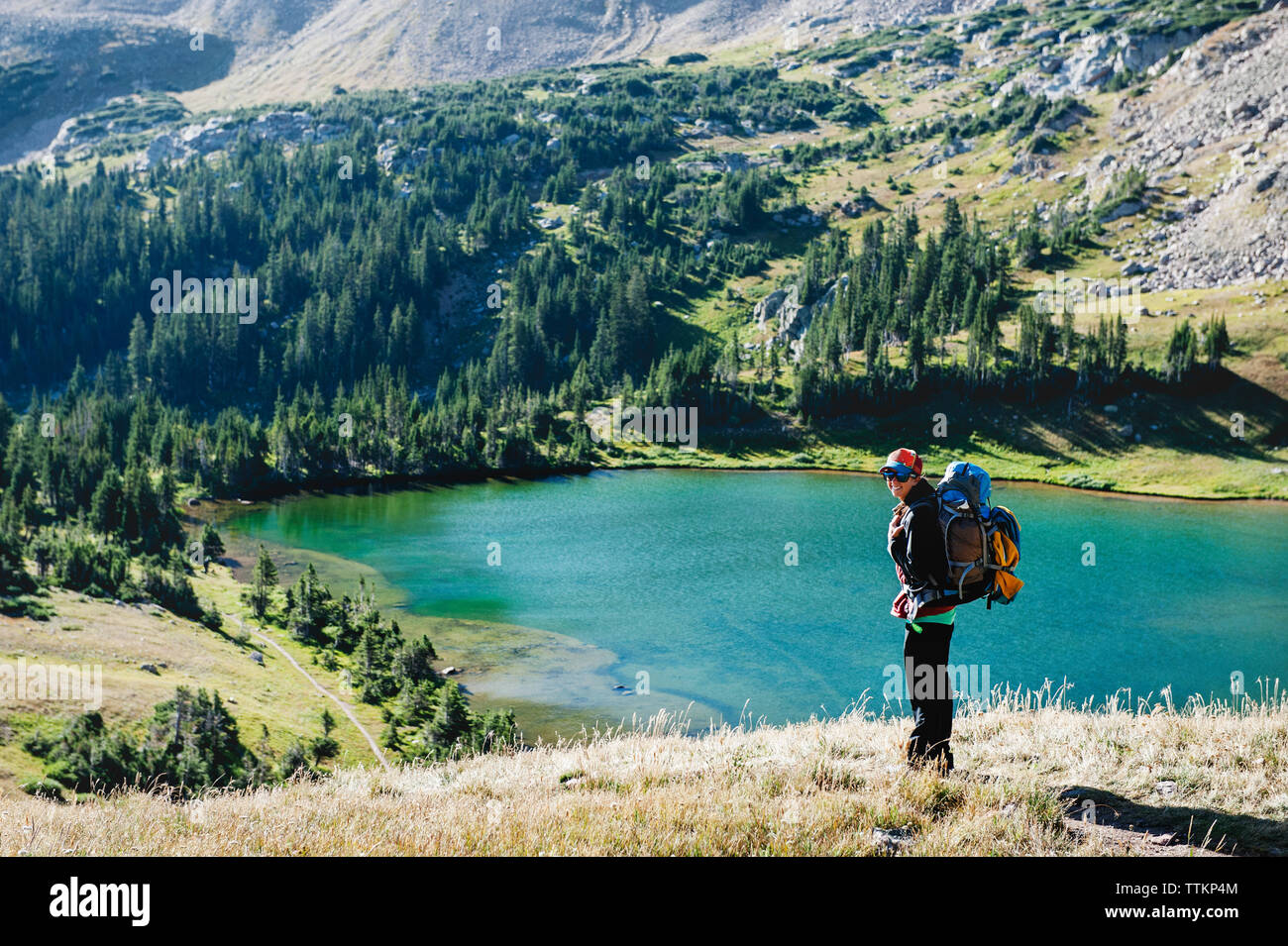 Donna sorridente che porta uno zaino in piedi contro il lago in montagna Foto Stock