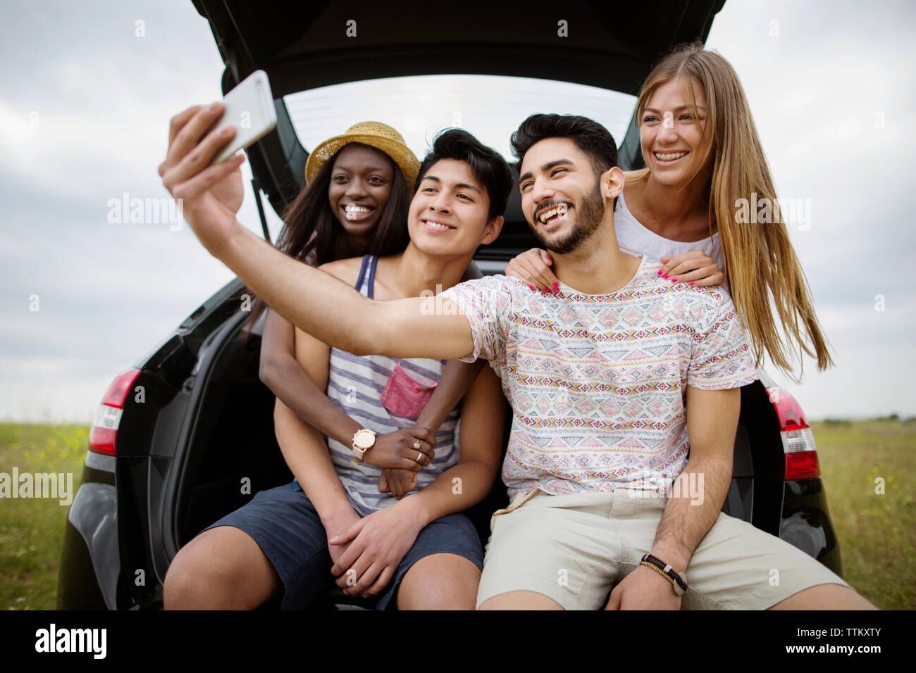 Felice multi-etnico amici prendendo selfie attraverso smart phone mentre sedendo nel baule auto sul campo Foto Stock