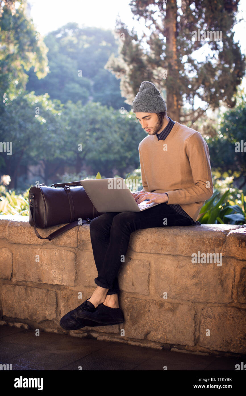 Uomo che utilizza il computer portatile mentre è seduto sulla parete di ritegno Foto Stock