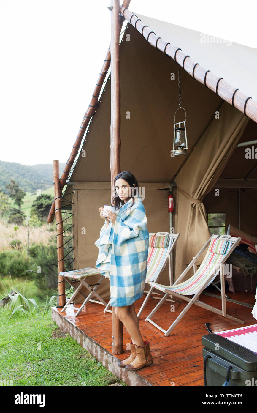 Donna avvolta in asciugamano di bere il caffè mentre si sta in piedi in tenda durante le vacanze Foto Stock