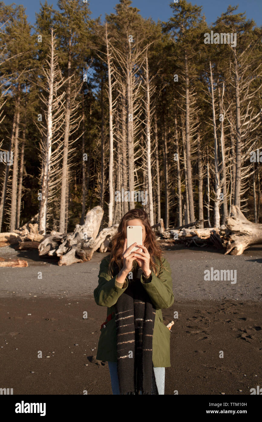 Giovane donna fotografando attraverso smart phone contro alberi sulla spiaggia Foto Stock