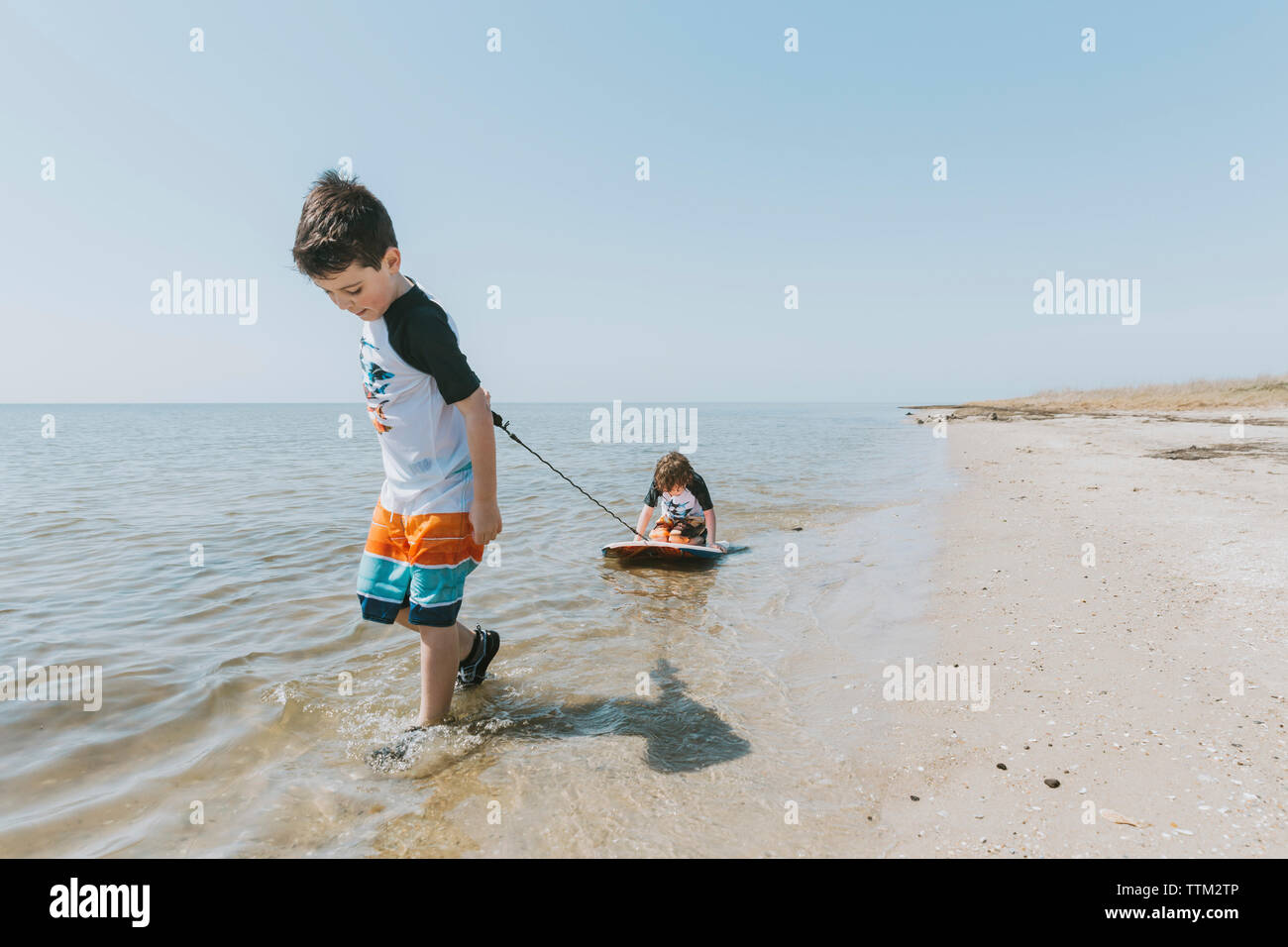 Ragazzo tirando il fratello seduto sulla tavola da surf in mare contro il cielo chiaro durante la giornata di sole Foto Stock