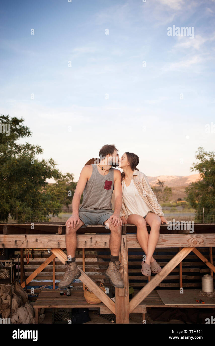 Affettuosa coppia di agricoltori kissing seduti sulla struttura in legno a farm contro sky Foto Stock