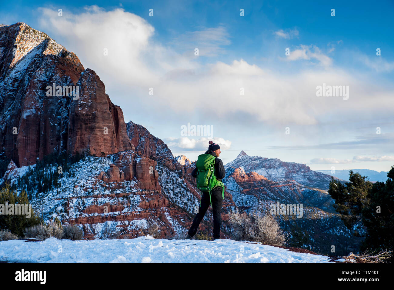 Vista posteriore dell'uomo con zaino verde permanente sulla neve in deserto contro formazioni di roccia e cielo nuvoloso durante il periodo invernale Foto Stock