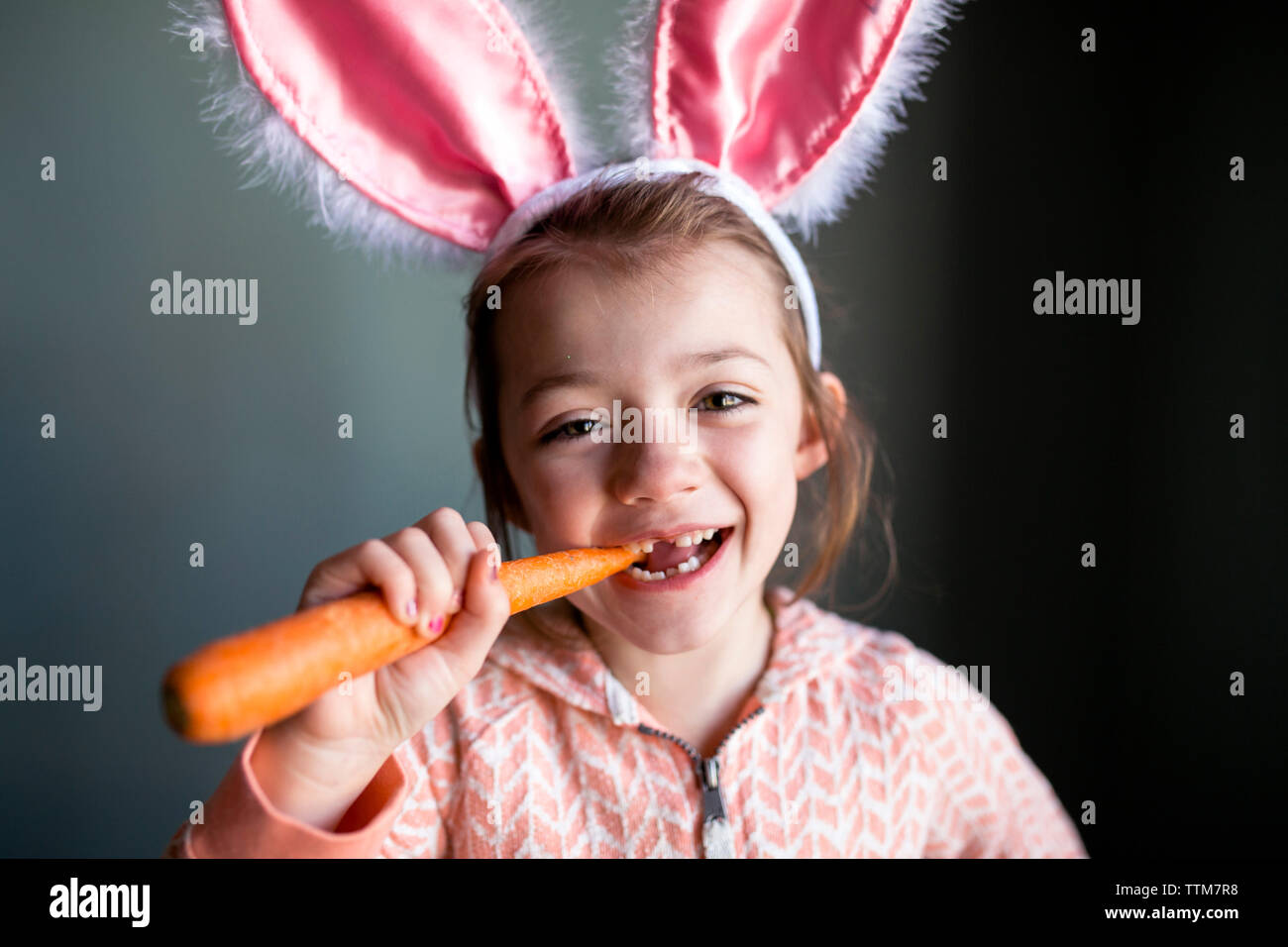 Ritratto di ragazza giocoso indossando orecchie di coniglio con archetto mentre morde la carota a casa Foto Stock