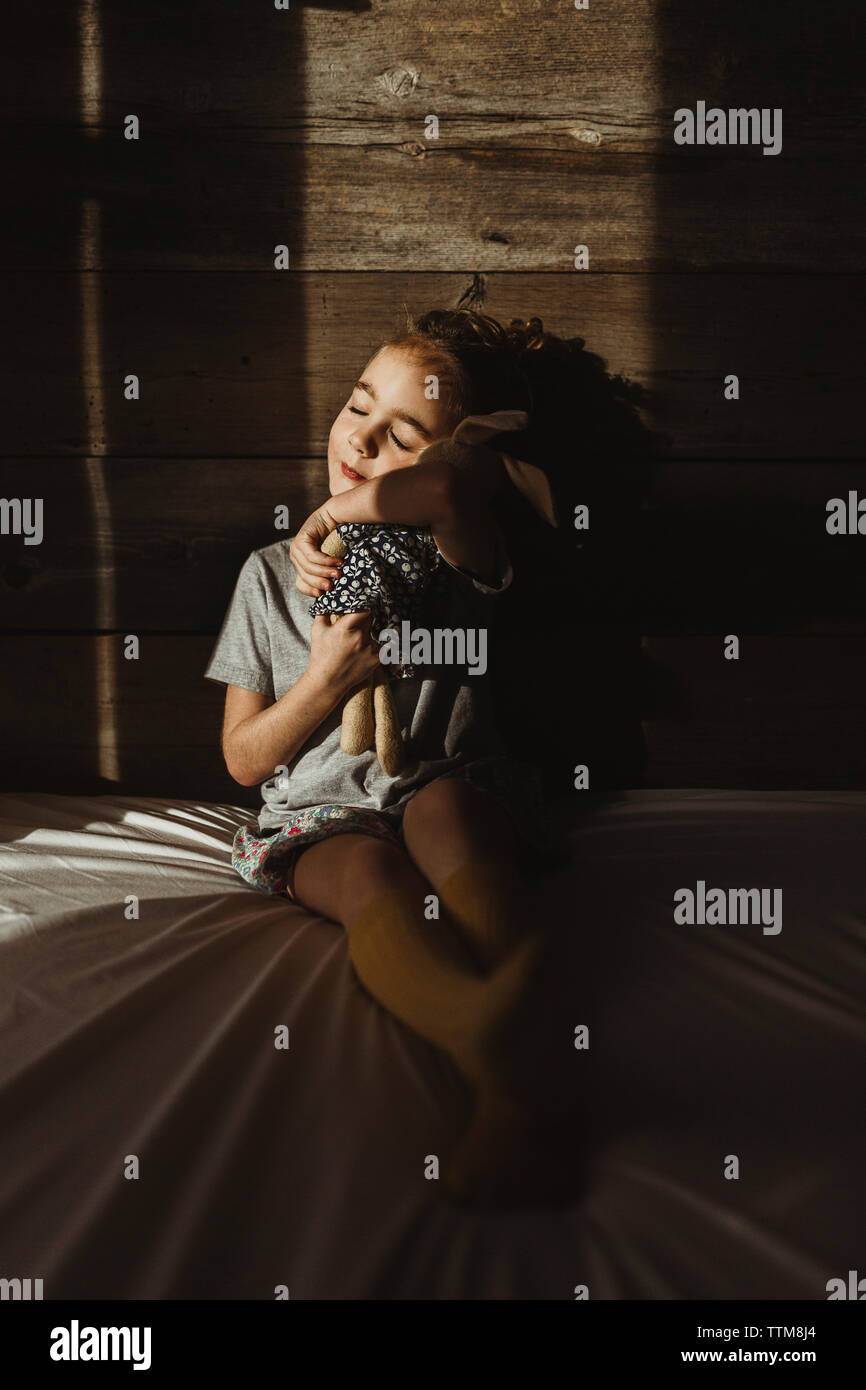 La ragazza con gli occhi chiusi abbracciando giocattolo imbottito seduti sul letto contro il legno nella camera oscura Foto Stock