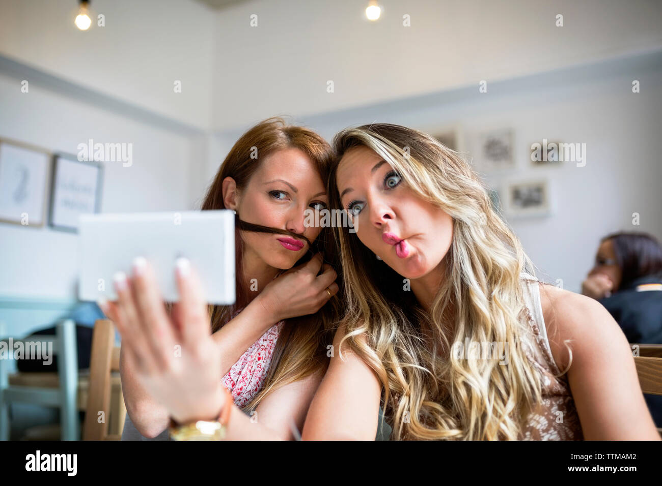 Amici di sesso femminile rendendo volti mentre prendendo selfie attraverso smart phone in cafe Foto Stock