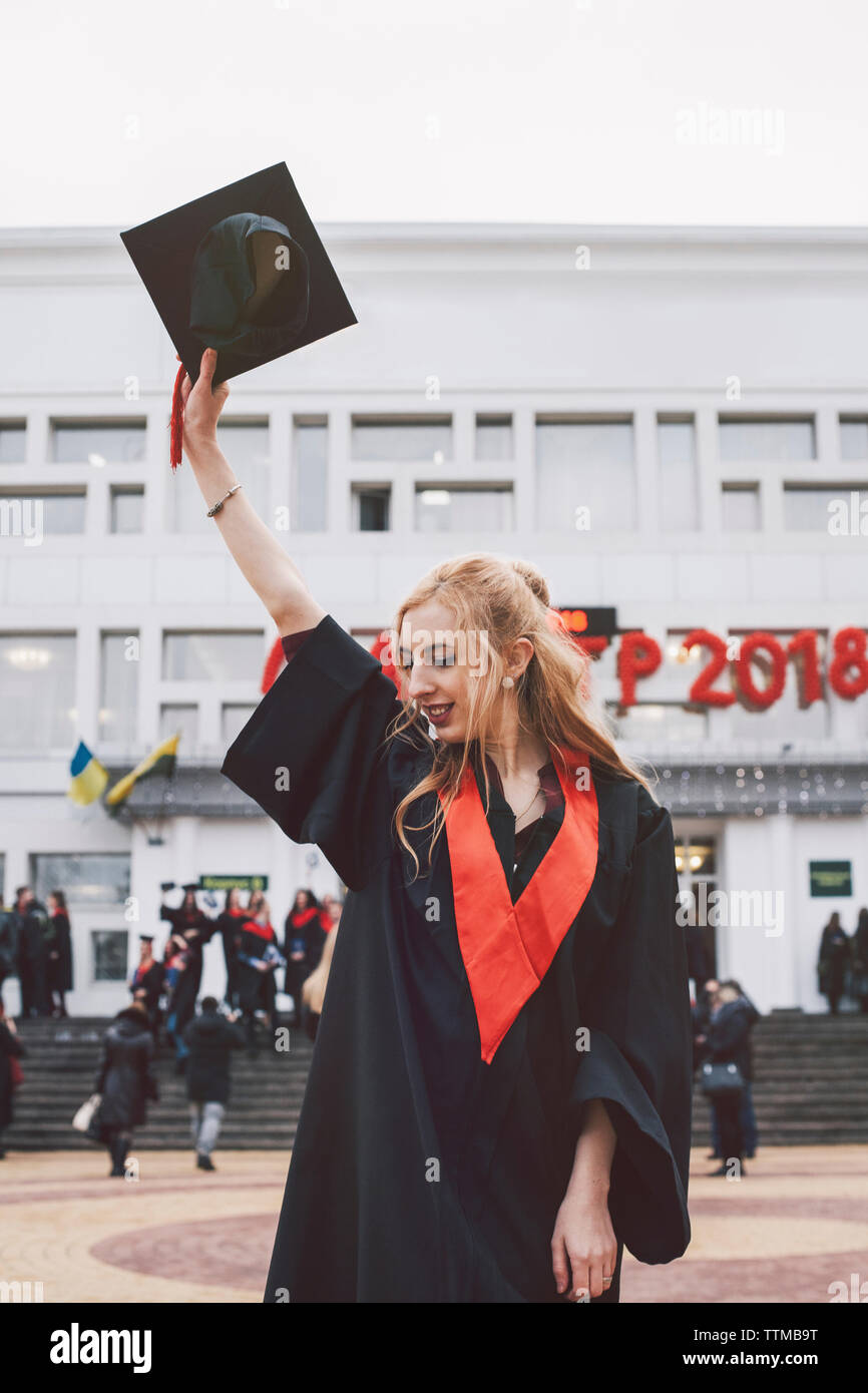 Donna in abito di graduazione holding mortarboard mentre in piedi contro la costruzione di Foto Stock