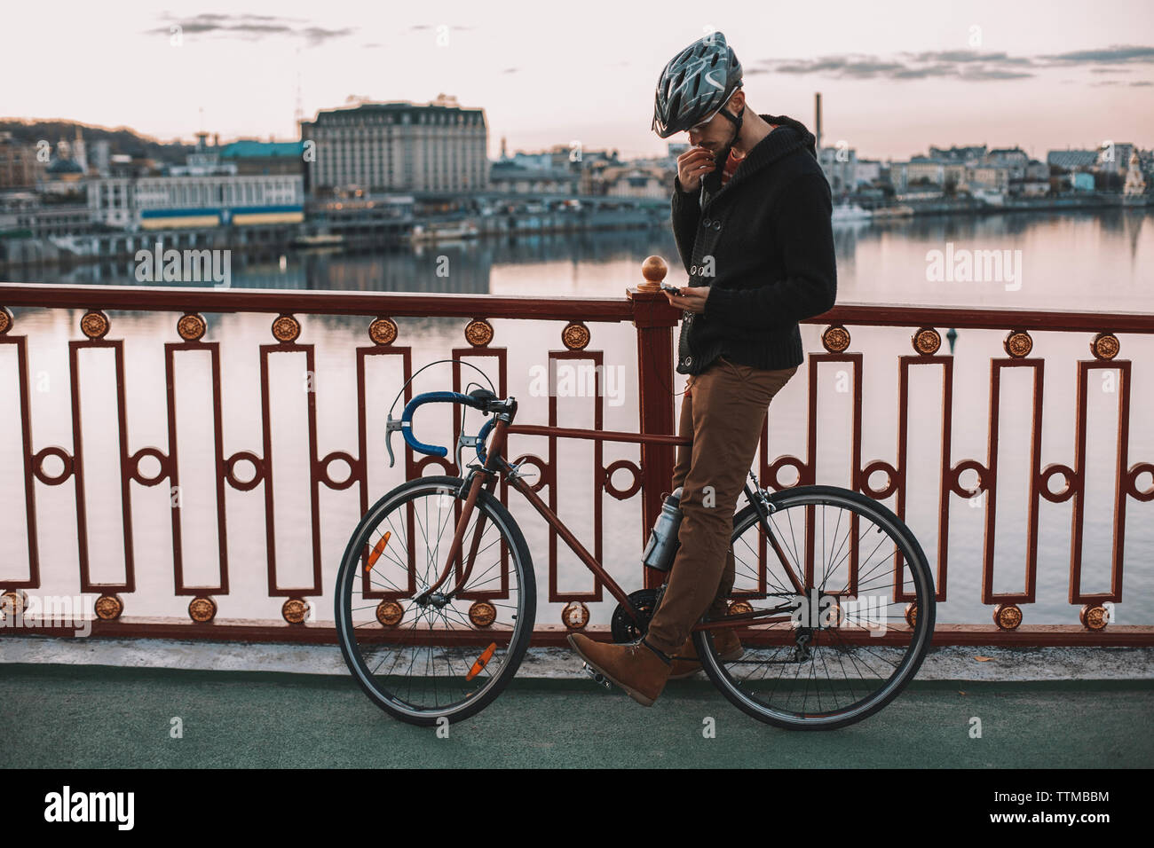 Ciclista utilizzando smart phone mentre in piedi con la bicicletta sul ponte sul fiume in città durante il tramonto Foto Stock