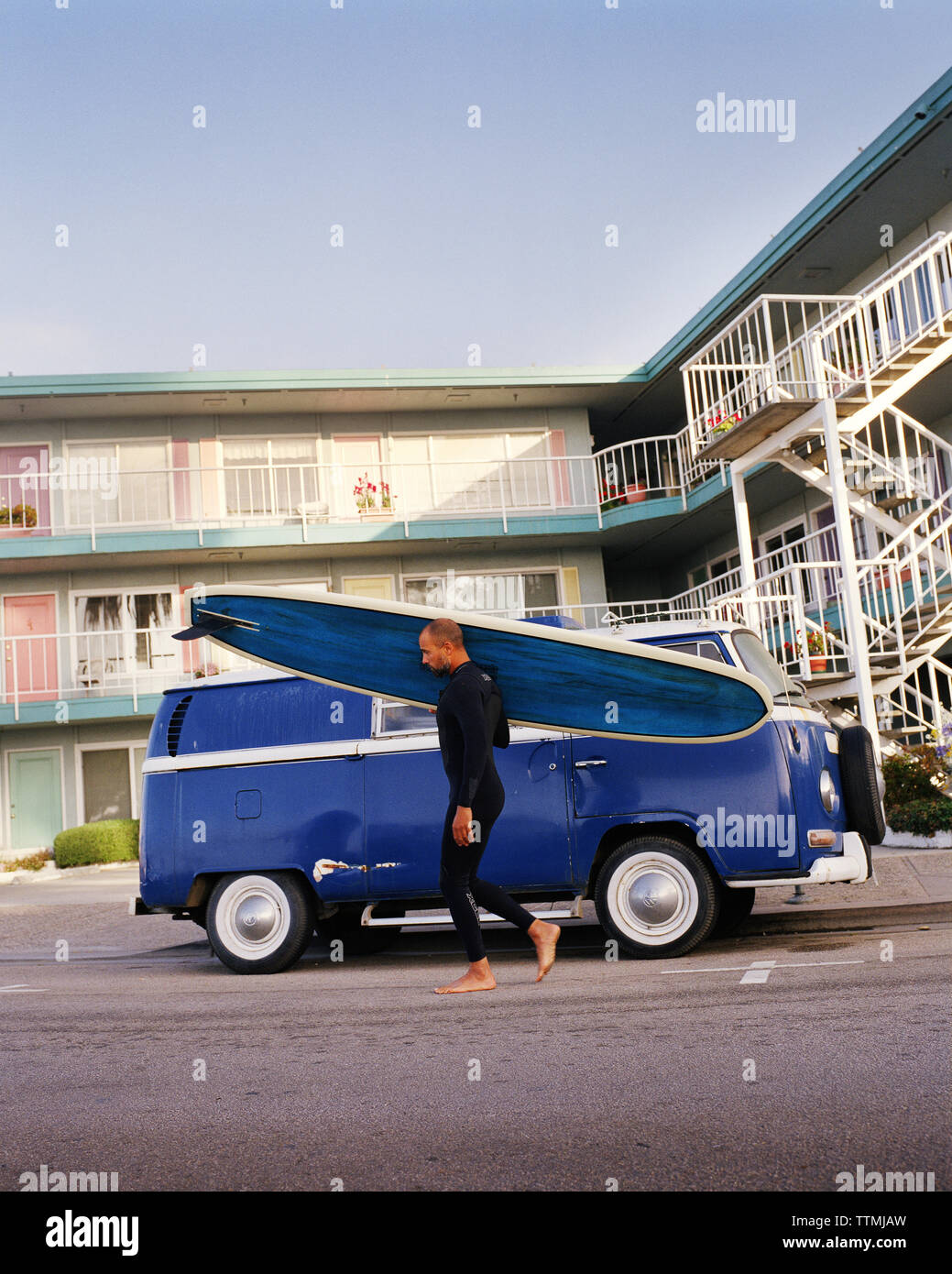 Stati Uniti, California, Capitola, un surfista con un blu longboard passeggiate in fronr di un blu bus VW sul suo modo di fare surf Foto Stock
