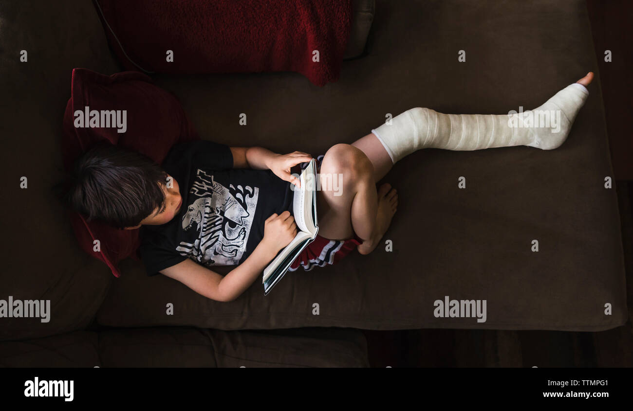 Angolo di Alta Vista del ragazzo con gamba rotta la lettura di libro mentre sdraiato sul divano di casa Foto Stock
