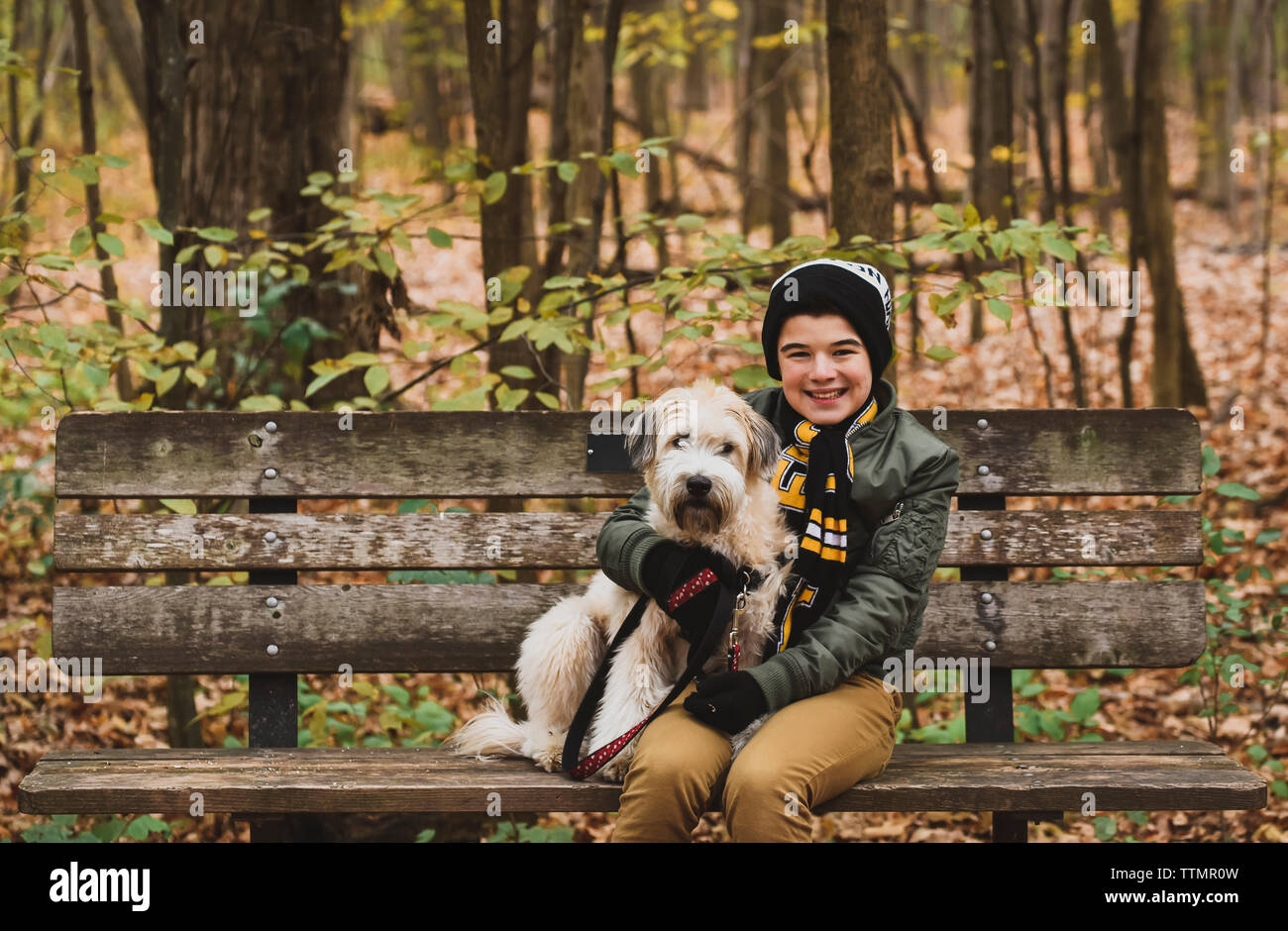 Ritratto di sorridere ragazzo seduto con il cane sul banco di prova nella foresta durante l'autunno Foto Stock