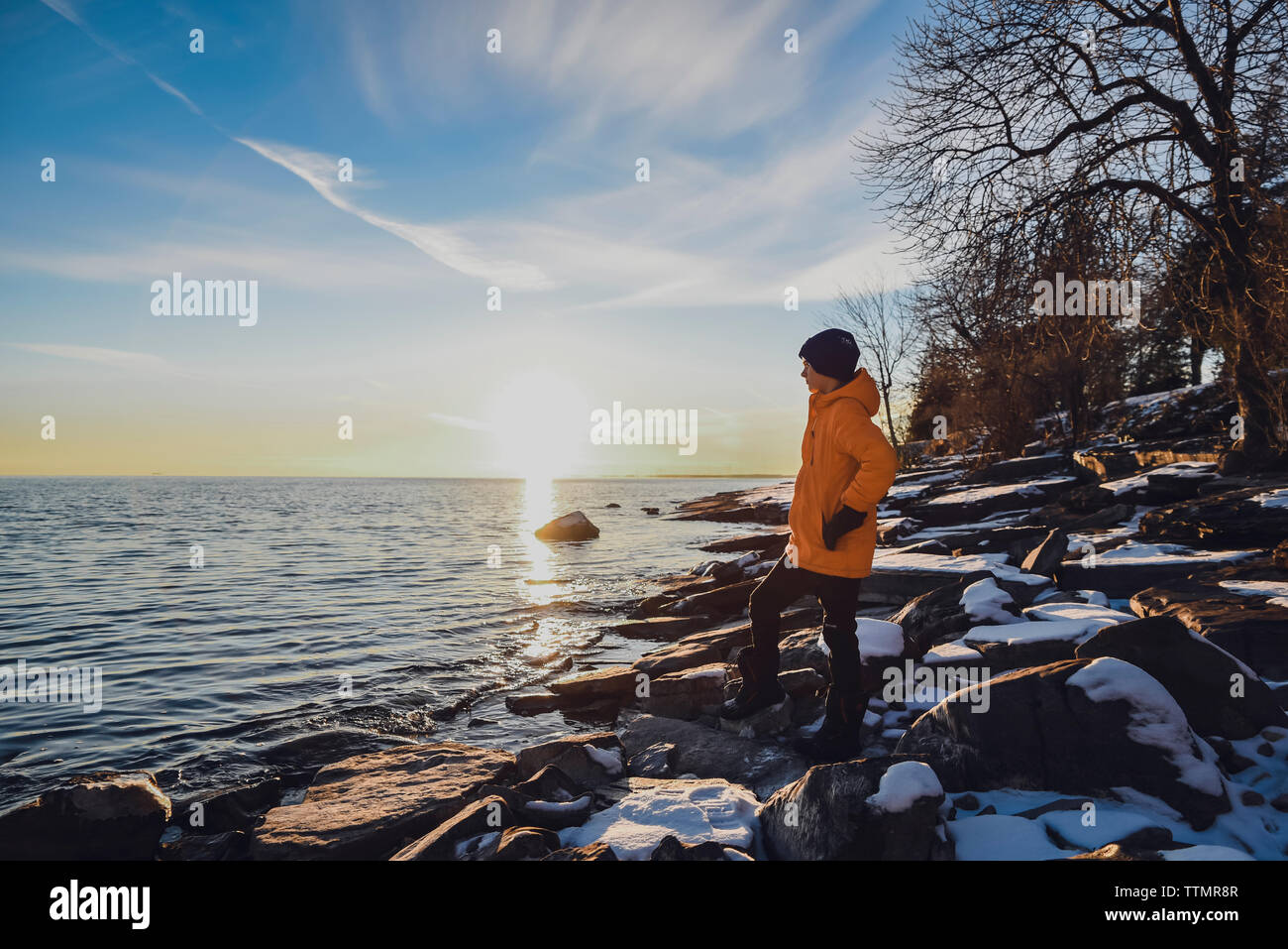 Ragazzo che sta da solo su rocce nevoso sul lungolago al tramonto in inverno. Foto Stock