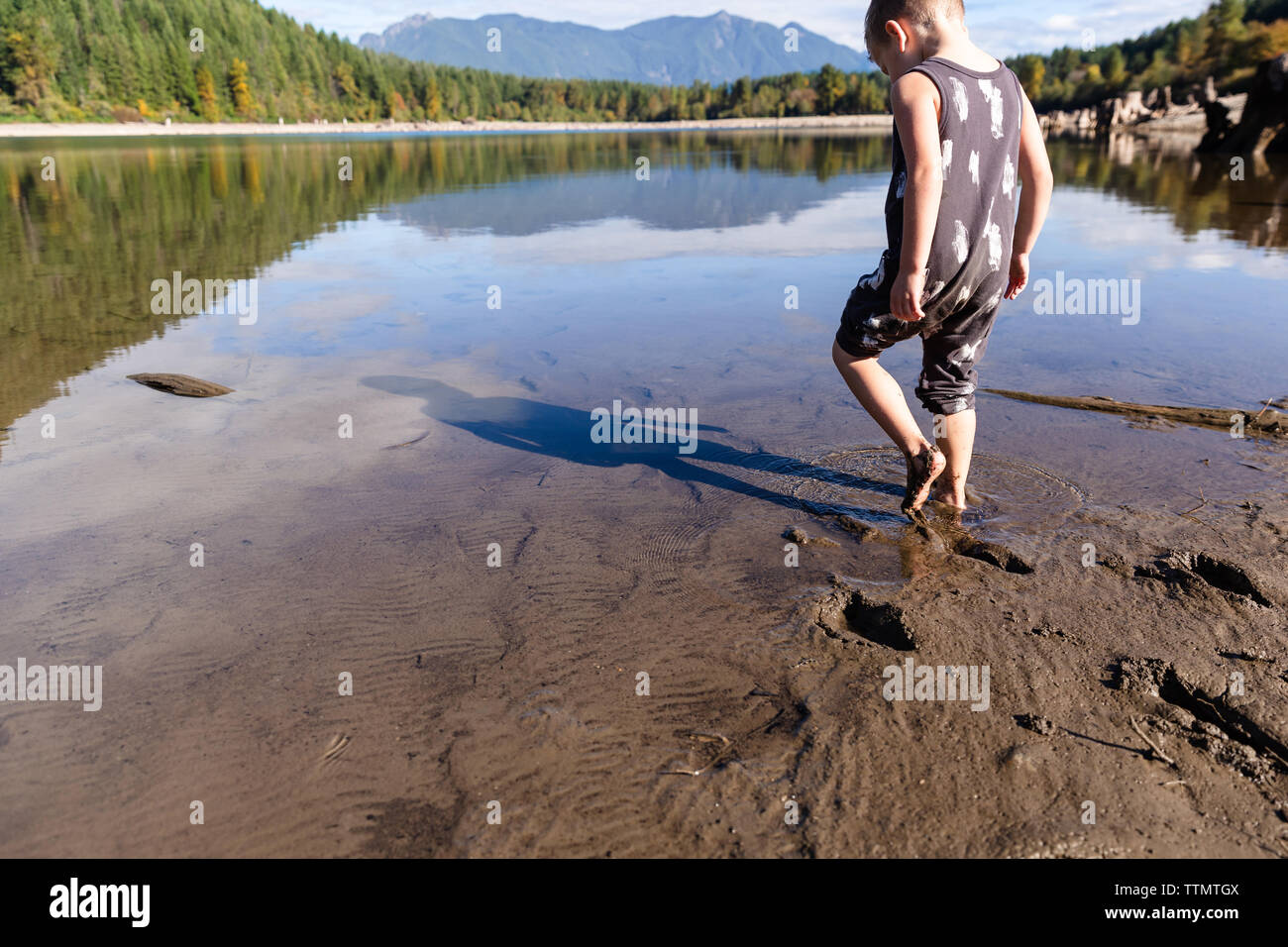 Un giovane bambino gioca a piedi nudi nel fango in prossimità di un lago sotto il sole Foto Stock