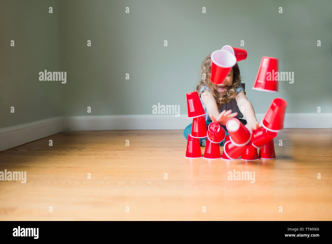 Giocoso ragazza piramide di rottura fatta di tazze monouso mentre è seduto sul pavimento a casa Foto Stock