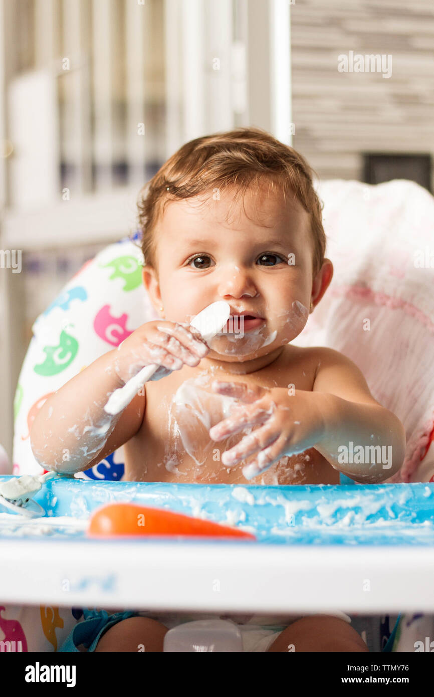 Baby colorate con yogurt mentre mangia yogurt nel suo seggiolone per mangiare Foto Stock