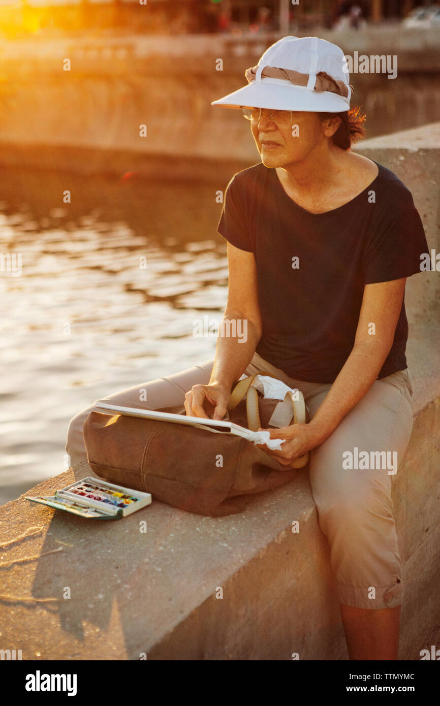 Senior donna dipinto su carta mentre è seduto alla parete di ritegno dal mare Foto Stock
