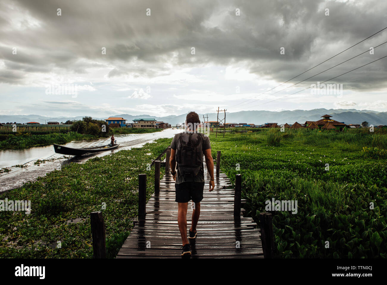 Vista posteriore dell'uomo con zaino camminando sul ponte pedonale dal Lago Inle contro il cielo nuvoloso Foto Stock