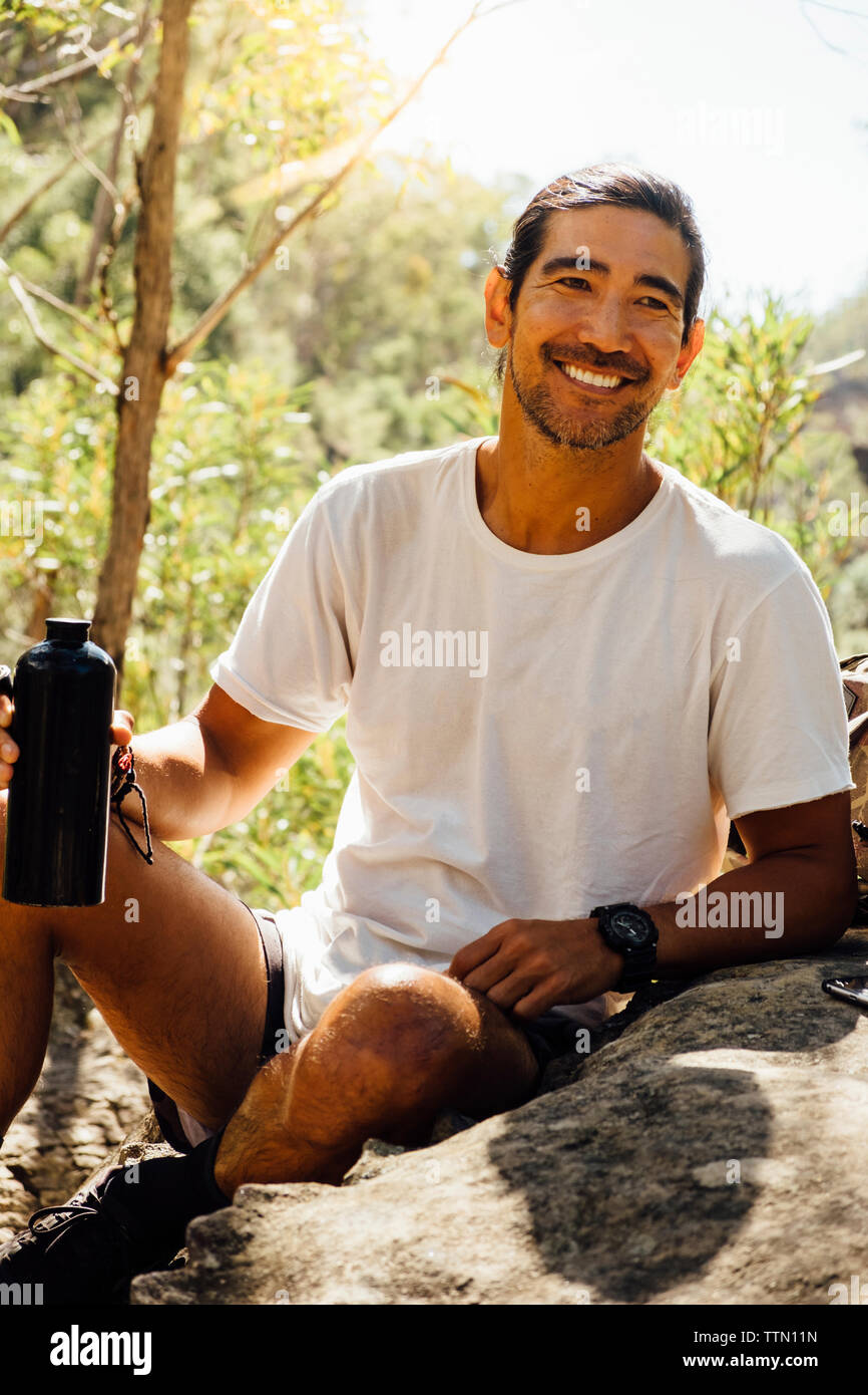 Felice escursionista maschio tenendo la bottiglia di acqua mentre è seduto sulla roccia contro gli alberi nelle foreste Foto Stock