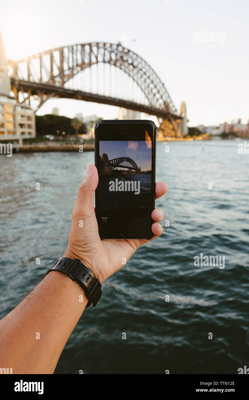 Ritagliate la mano dell'uomo a fotografare il Ponte del Porto di Sydney su Port Jackson contro il cielo chiaro in città Foto Stock