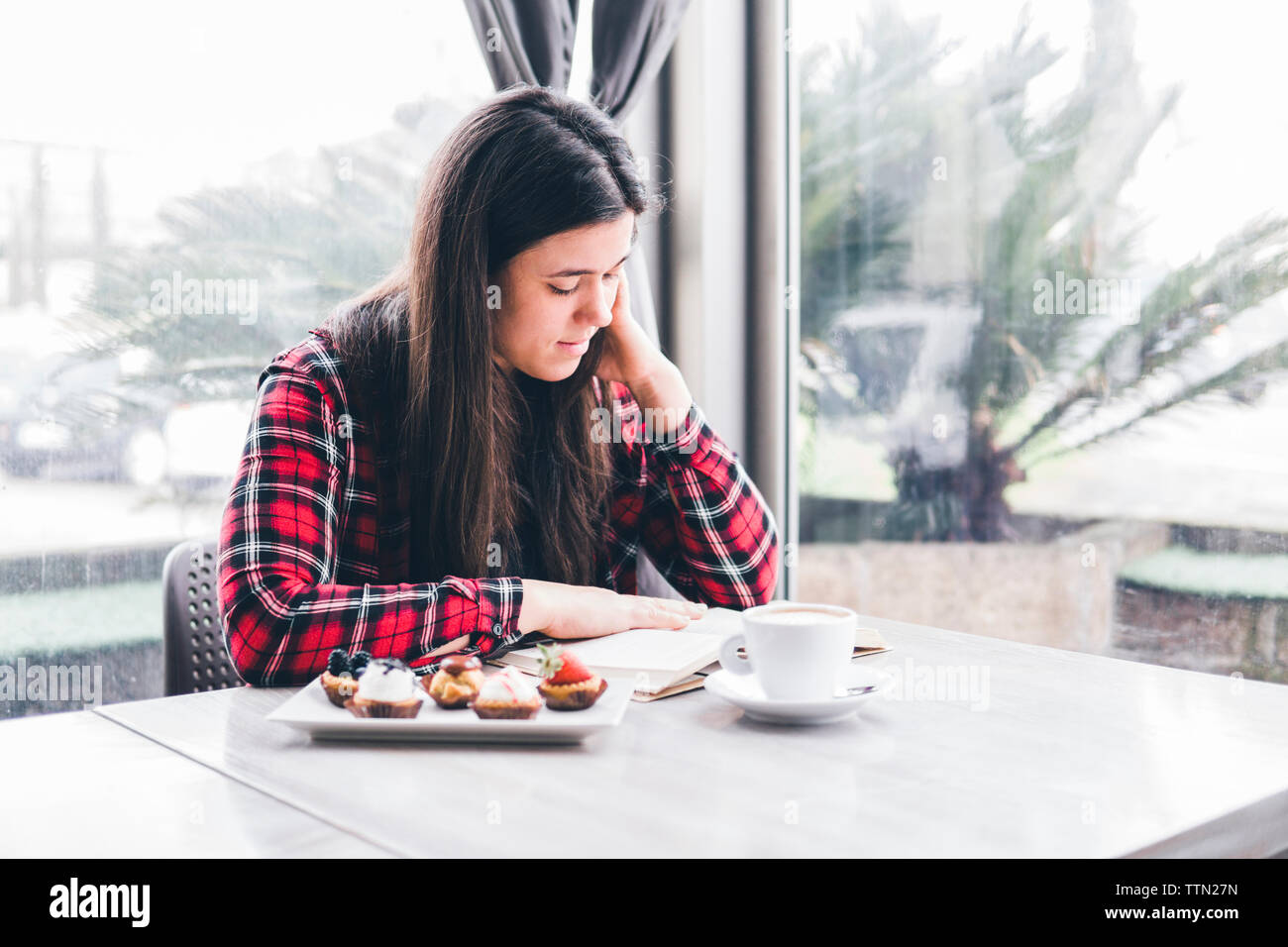 Donna con cibi dolci e caffè sulla tabella Rubrica lettura mentre è seduto da windows in cafe Foto Stock