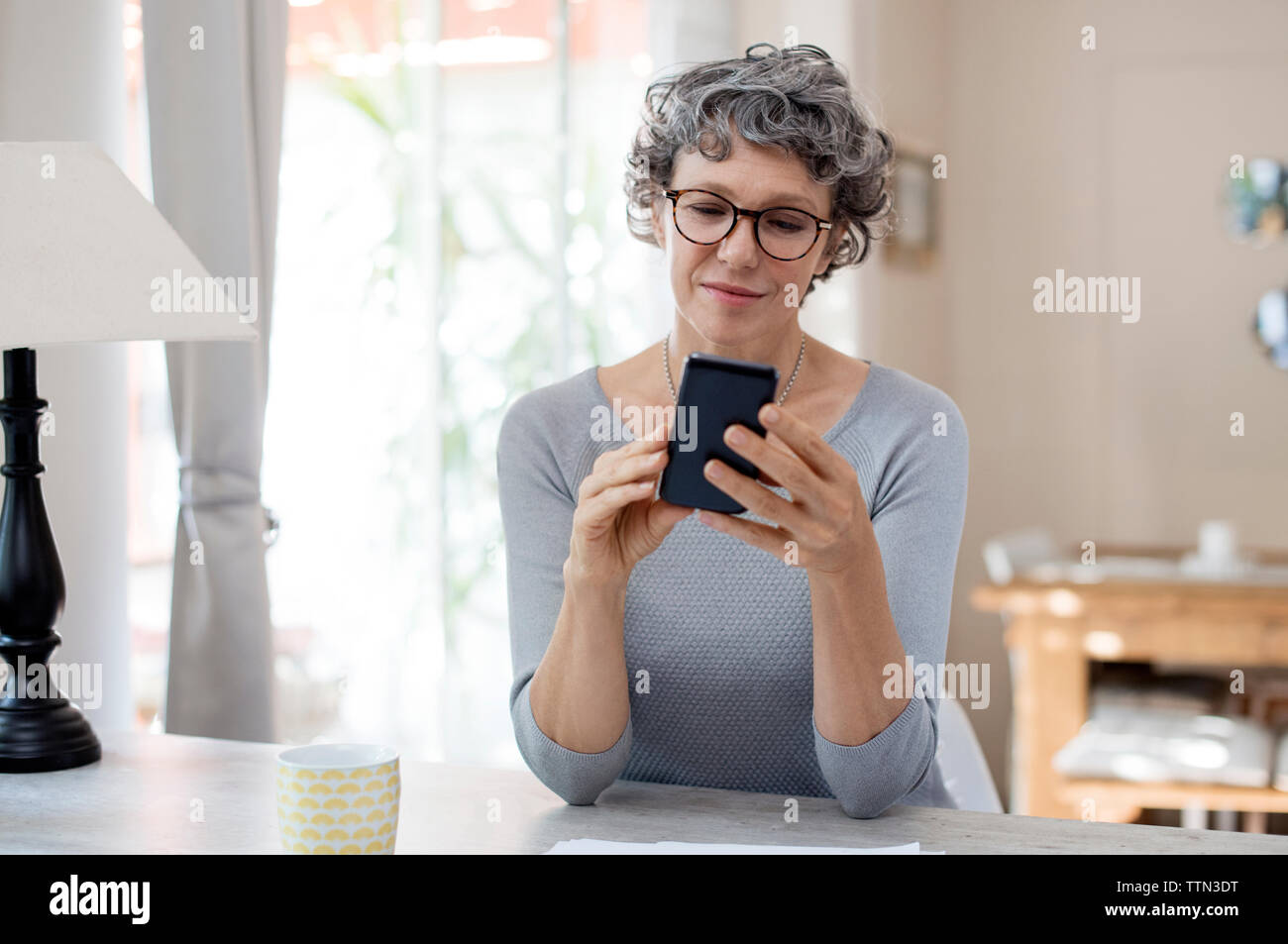 Donna matura utilizzando smart phone sul tavolo a casa Foto Stock
