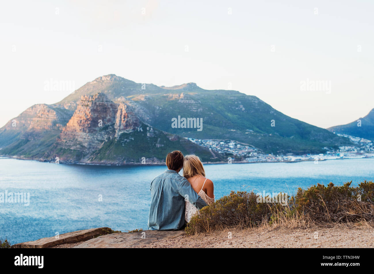 Vista posteriore del giovane seduto sulla collina contro il mare e montagna rocciosa Foto Stock