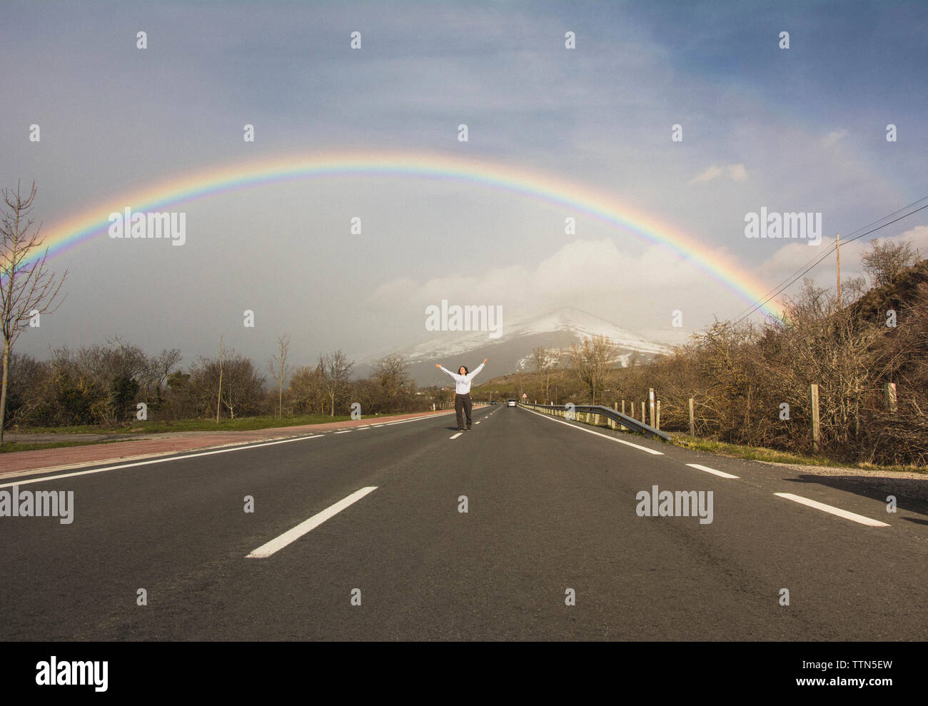 A metà distanza vista della giovane donna con le braccia sollevate in piedi sulla strada contro arcobaleno e cielo nuvoloso durante il periodo invernale Foto Stock