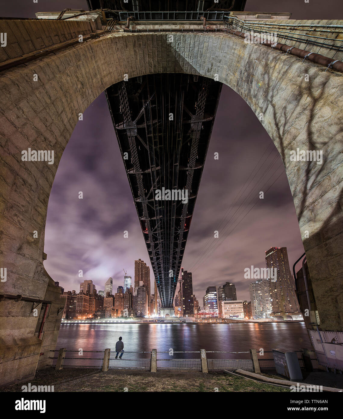 Uomo seduto sulla ringhiera sotto il ponte sul fiume e la città illuminata al crepuscolo Foto Stock