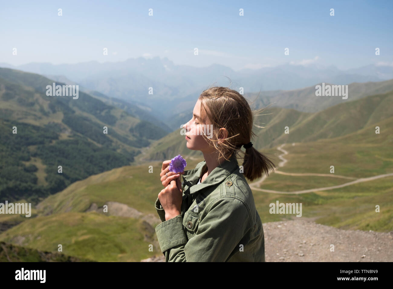 Vista laterale della donna con gli occhi chiusi fiore di contenimento mentre si sta in piedi sul monte contro sky durante la giornata di sole Foto Stock