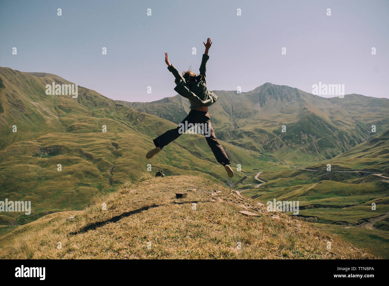 Vista posteriore della donna felice con le braccia sollevate il salto sulla montagna contro il cielo chiaro durante la giornata di sole Foto Stock