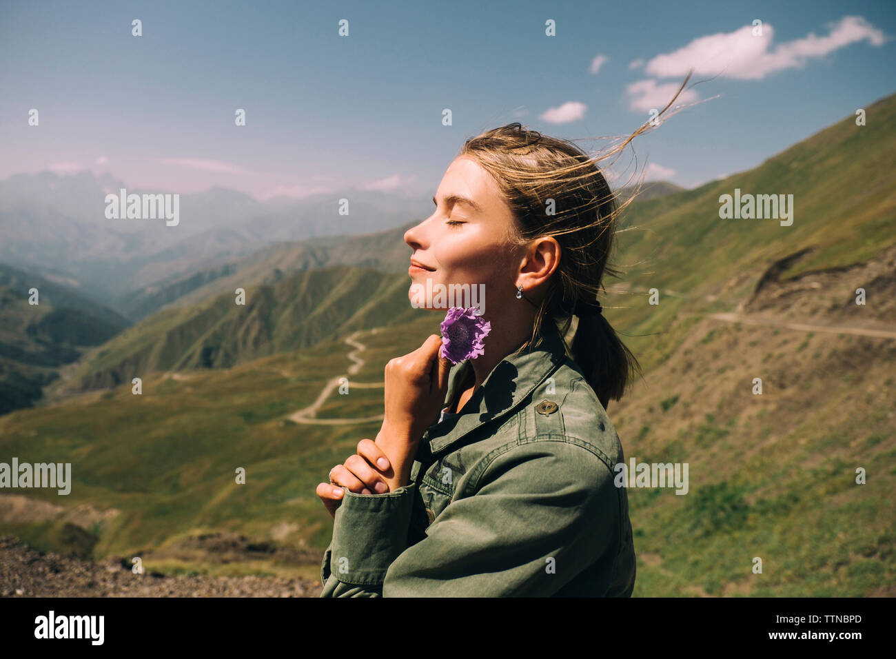 Vista laterale della donna sorridente con gli occhi chiusi fiore di contenimento mentre si sta in piedi sul monte contro sky durante la giornata di sole Foto Stock